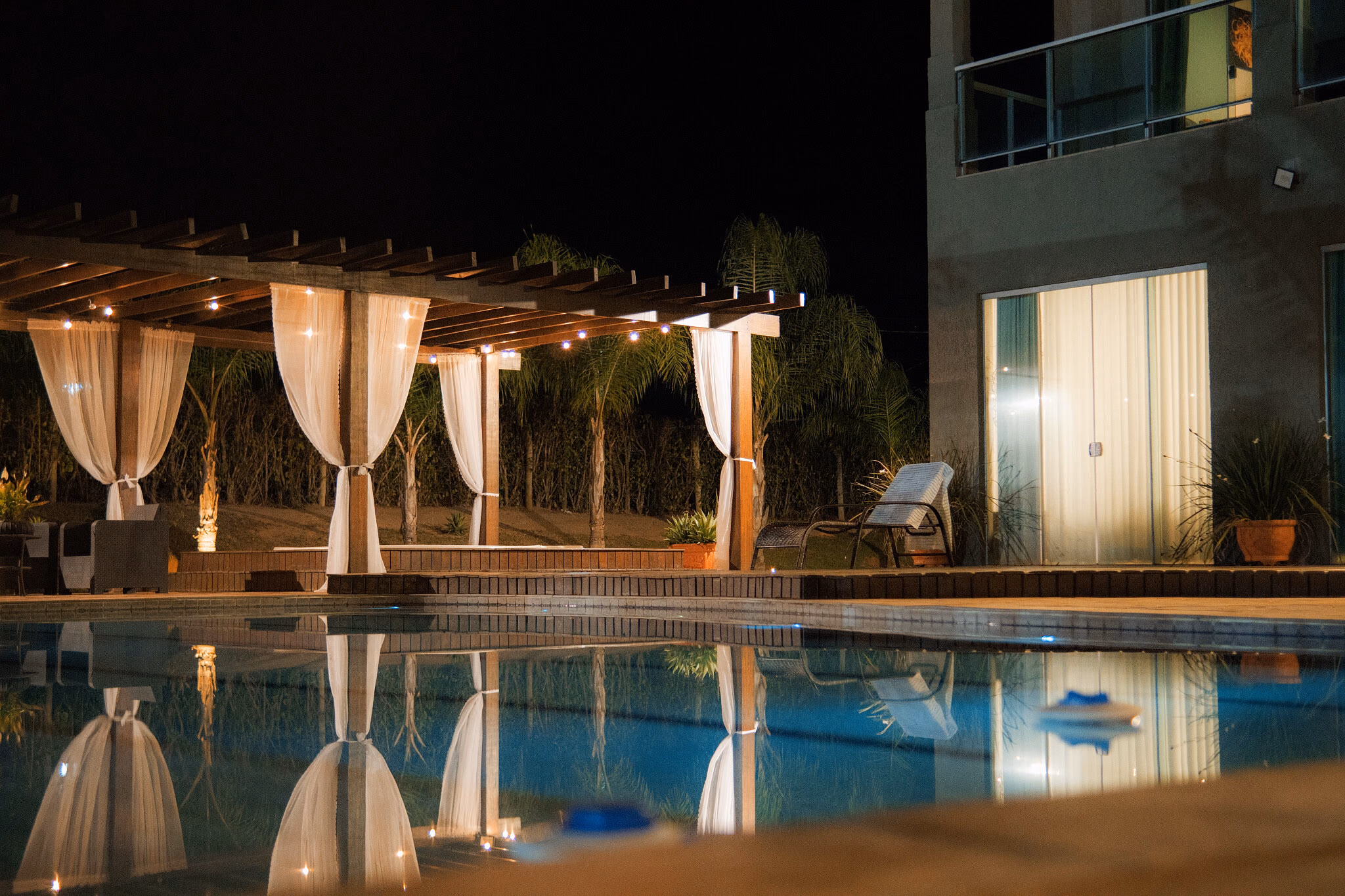 Mol Residence ao entardecer — piscina e pergolado iluminados, fachada da casa refletida na água