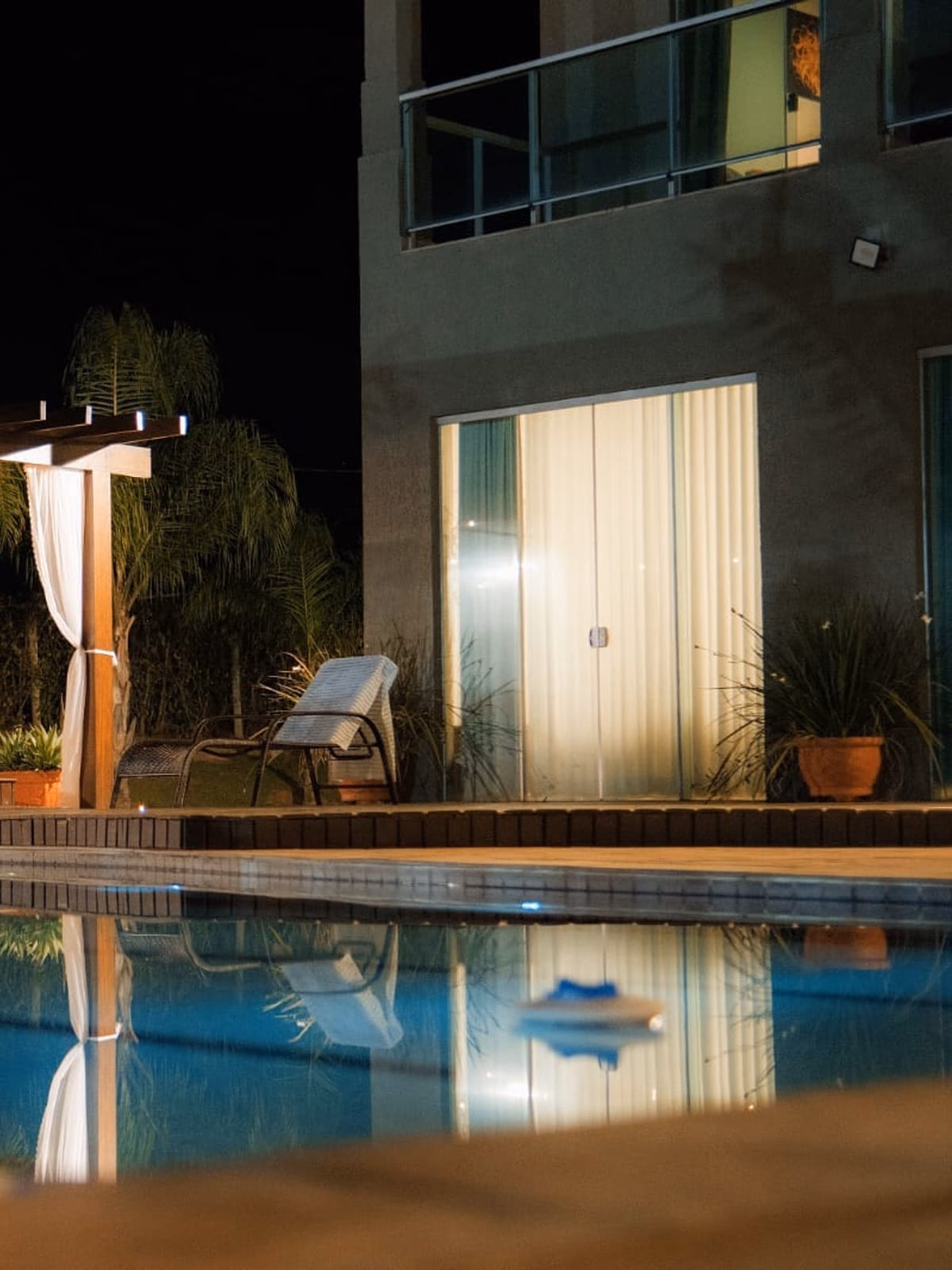 Pergolado com cortinas brancas à noite, reflexo na piscina